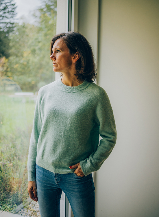 Eine Frau steht in ruhiger Pose am Fenster und blickt nachdenklich nach draußen
