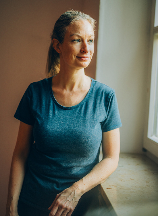 Frau in blauem T-Shirt schaut umgeben von sanftem Tageslicht leicht lächelnd aus dem Fenster