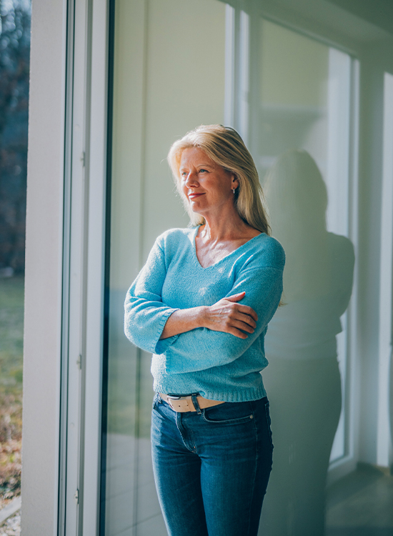 Eine Frau in einem blauen Pulli steht mit verschränkten Armen an einer geöffneten Glastür und schaut nachdenklich nach draußen